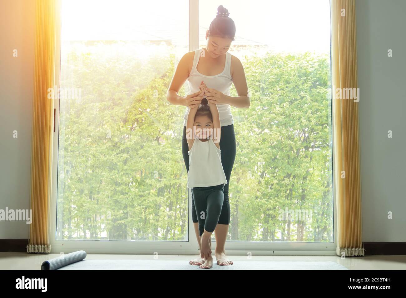 Mamma asiatica pratica yoga con una figlia adorabile a casa, cercare di insegnare la figlia giovane a fare yoga posa albero o vrksasana, mentre l'esercizio a casa. Foto Stock