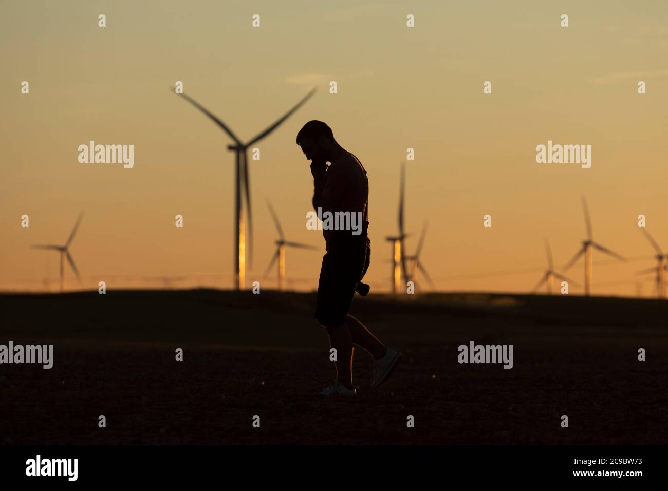Un uomo cammina parlando al telefono in campagna al tramonto, dopo aver scattato foto di turbine eoliche lontane, all'orizzonte. Foto Stock