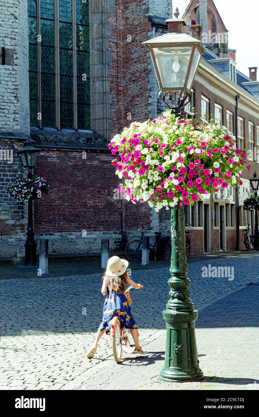 Leiden, Paesi Bassi - 22 luglio 2020: Bambina con cappello soleggiato su una bicicletta che aspetta accanto a una storica strada con fiori colorati Foto Stock