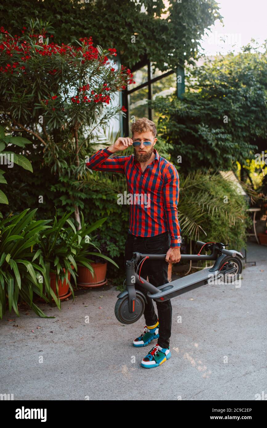 Un bel giovane uomo caucasico con gli occhiali da sole è in piedi in strada con scooter Foto Stock