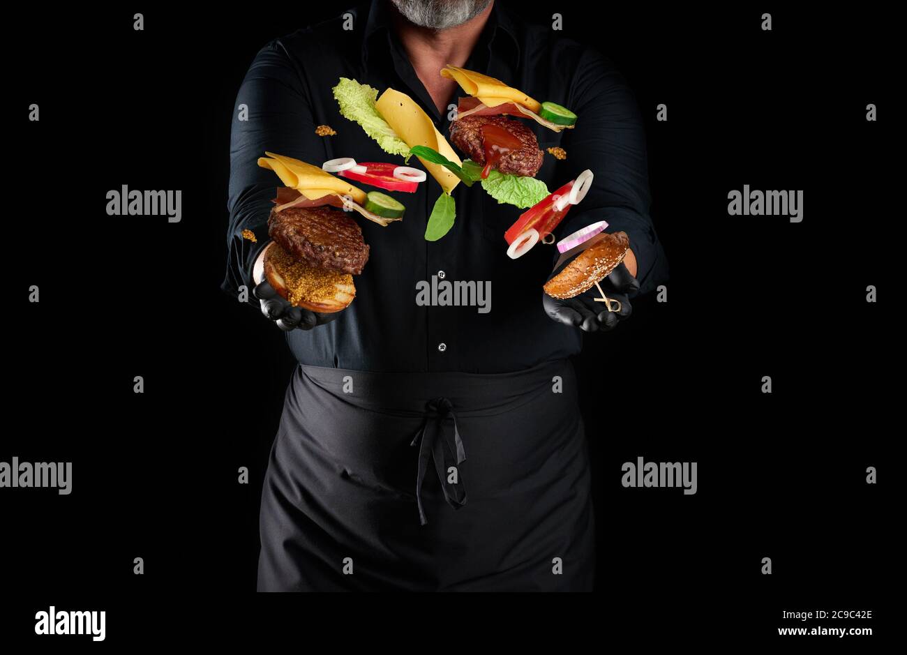 chef in una camicia nera, grembiule e guanti neri in lattice si trova su uno sfondo nero, in mani battenti cheeseburger ingredienti: un mordere con semi di sesamo Foto Stock