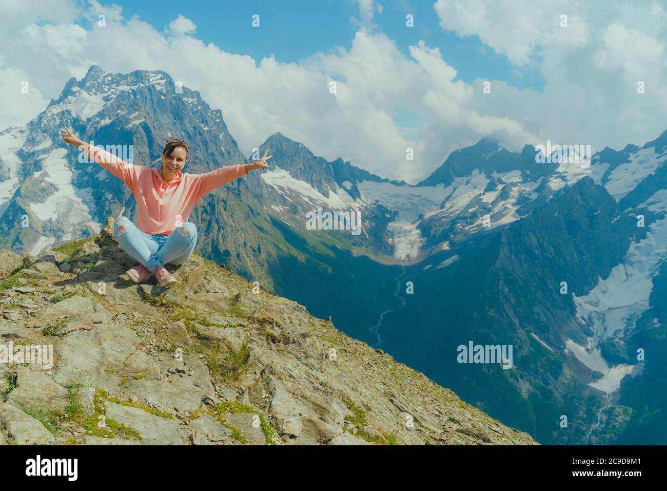 Giovane donna felice seduta sulla roccia contro il cielo nuvoloso in una giornata di sole in montagna. Il viaggiatore femminile ammira una splendida vista nella zona montagnosa. Foto Stock