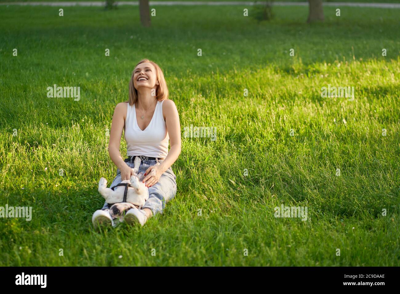 Giovane donna ridente seduta sull'erba con il bulldog francese maschio sulle gambe, divertendosi. Vista frontale della splendida caucasica caucasica ragazza caucasica godendo di una calda giornata estiva con il cane, accarezzando il ventre nel parco della città. Foto Stock