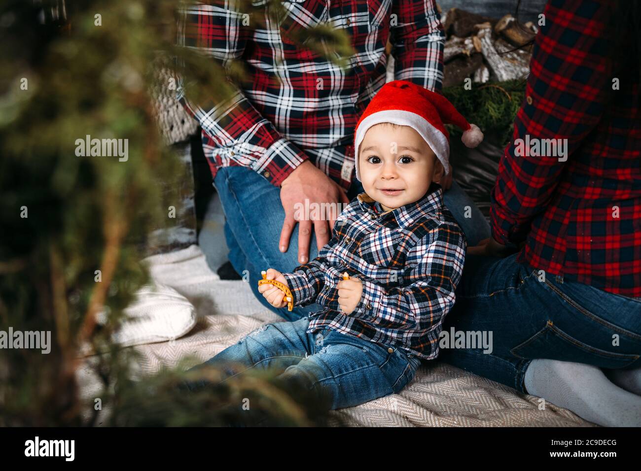 Ritratto di famiglia di Natale di giovani felici genitori sorridenti che giocano con il piccolo capretto in cappello rosso di santa vicino all'albero di natale. Vacanze invernali Natale e nuovo Foto Stock