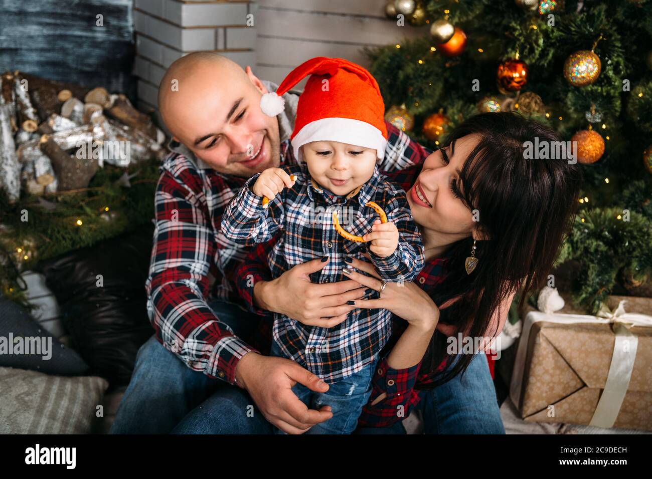 Ritratto di famiglia di Natale di giovani felici genitori sorridenti che giocano con il piccolo capretto in cappello rosso di santa vicino all'albero di natale. Vacanze invernali Natale e nuovo Foto Stock