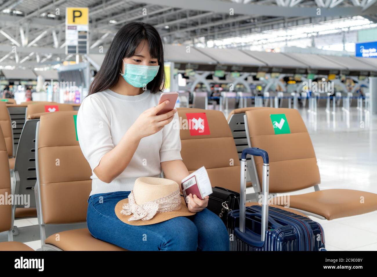 Asian donna turistica indossare maschera utilizzando il telefono cellulare ricerca dello stato del volo della compagnia aerea e seduta sociale sedia di distanza in aeroporto terminal durante pro Foto Stock