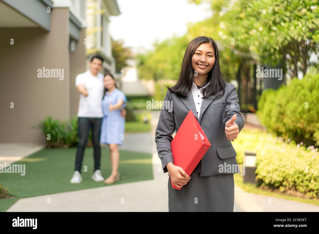 Agente immobiliare asiatico o donna immobiliare sorridente e tenendo file rosso con il pollice su con felice coppia asiatica abbracciando di fronte a casa tua. Foto Stock