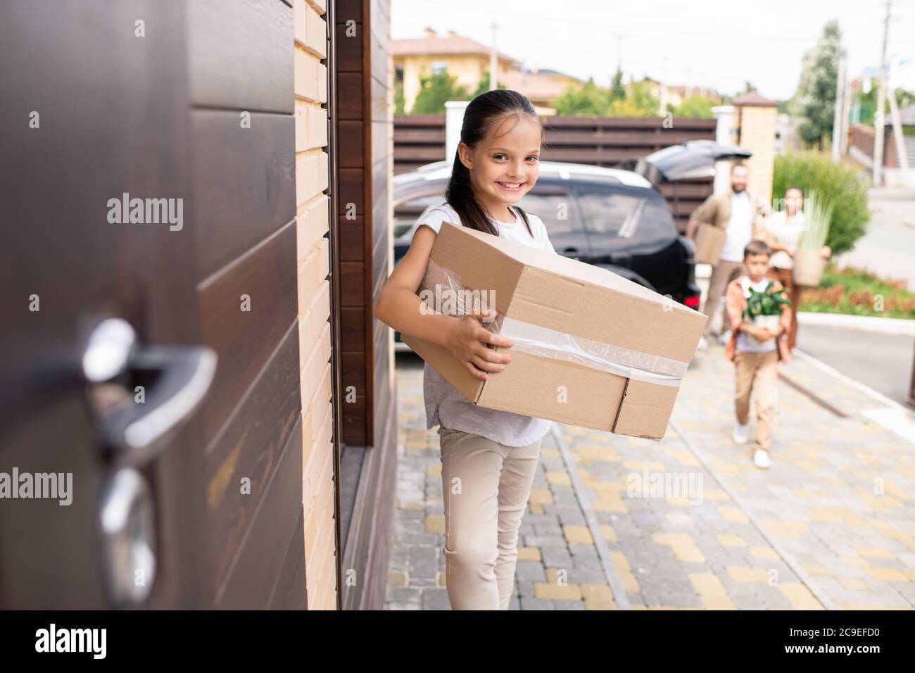 Ritratto di felice bella ragazza con ponytail che tiene grande scatola in movimento e in piedi alla porta di nuova casa Foto Stock