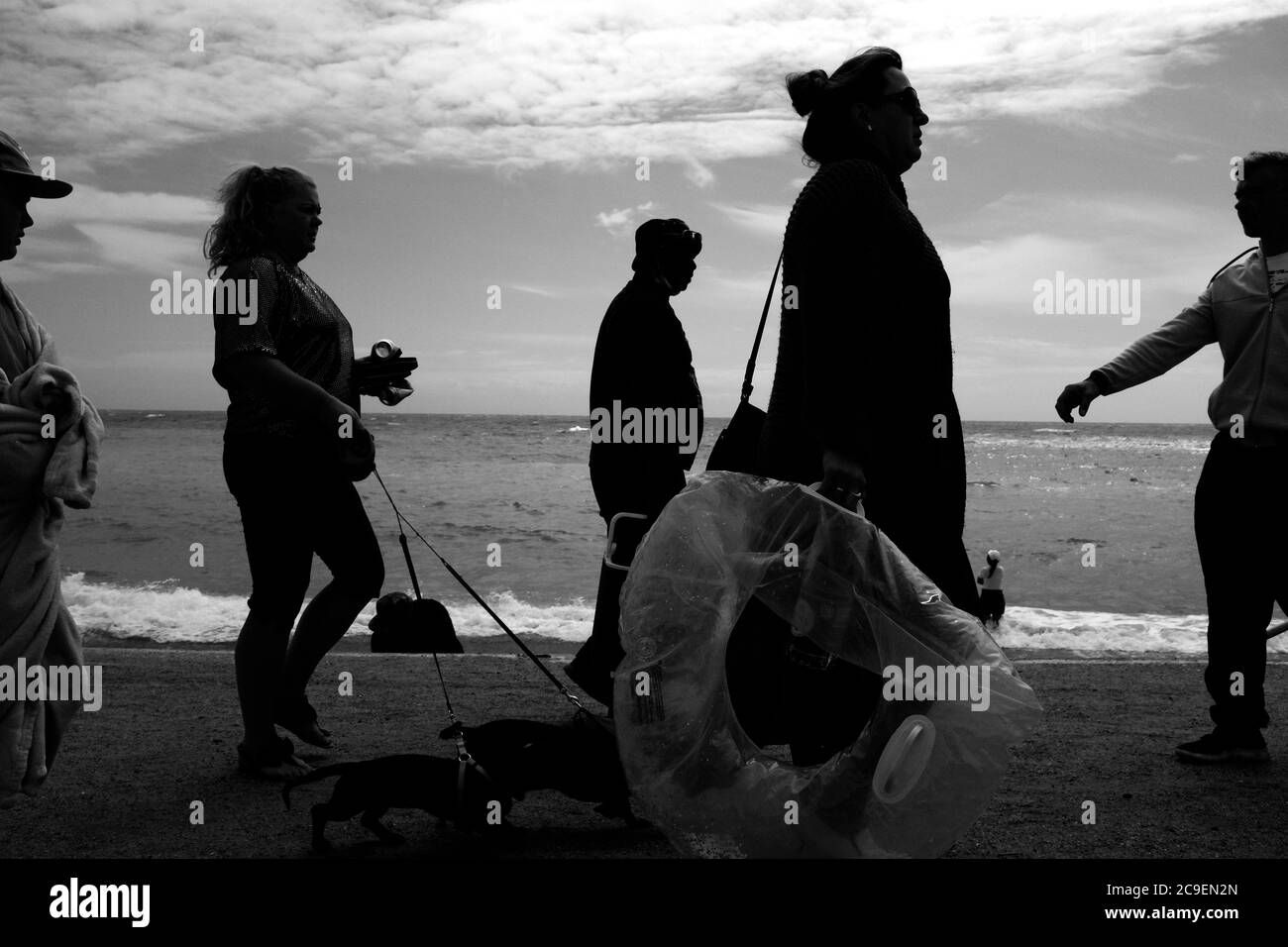 I beachgoers che si prosteggiano lungo il muro di mare di revetment in silhouette completamente vestito camminando i cani a Freshwater Bay sull'Isola di Wight. Foto Stock
