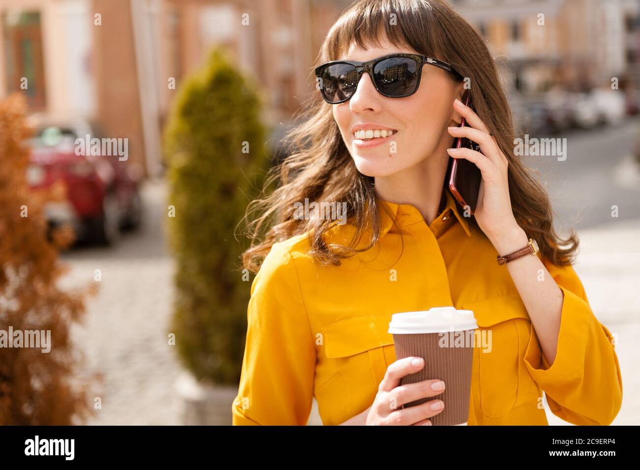 Happy girl in camicia gialla cammina per la strada, beve caffè e parla al telefono. Foto Stock