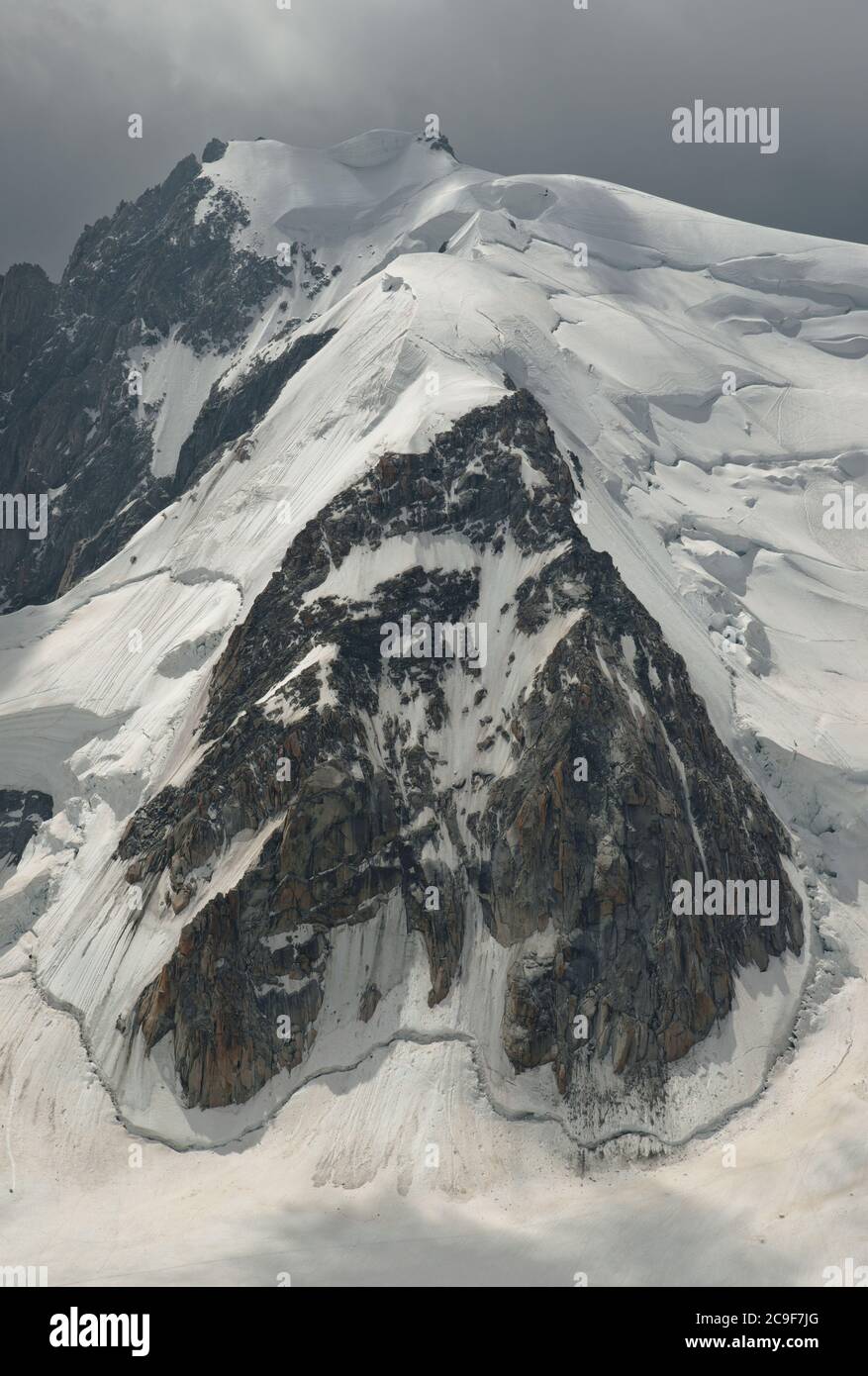 Parete rocciosa di forma insolitamente sul lato di una montagna innevata. La luce brillante si illumina attraverso un cielo nuvoloso sopra. Composizione grafica. Foto Stock