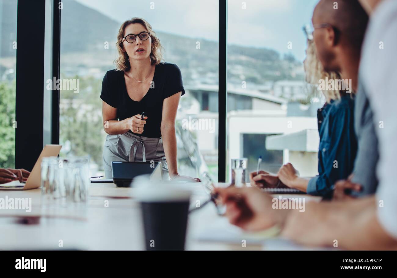 donna d'affari che si rivolge a una riunione in sala riunioni con colleghi seduti al tavolo. Manager donna che si rivolge al suo team durante una riunione. Foto Stock