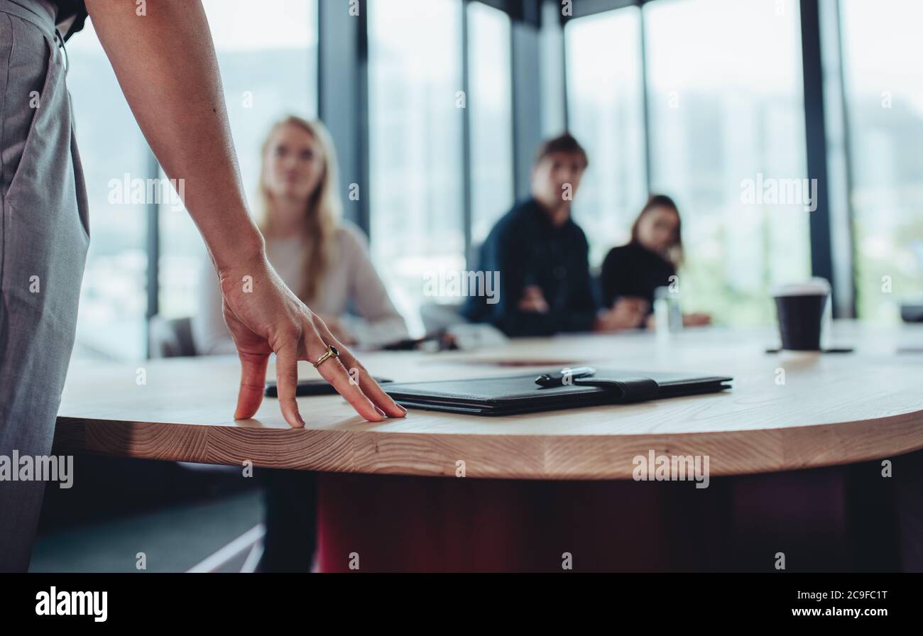 Donna manager in piedi al tavolo e rivolgendosi al suo team seduto nella sala riunioni. Mettere a fuoco sulla mano della donna d'affari in tavola mentre parla con il collega Foto Stock