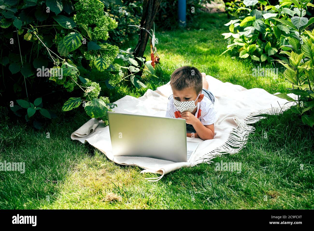 Ragazzino in una maschera, sdraiato su una plaid sull'erba con un laptop durante il periodo di quarantena. Foto Stock