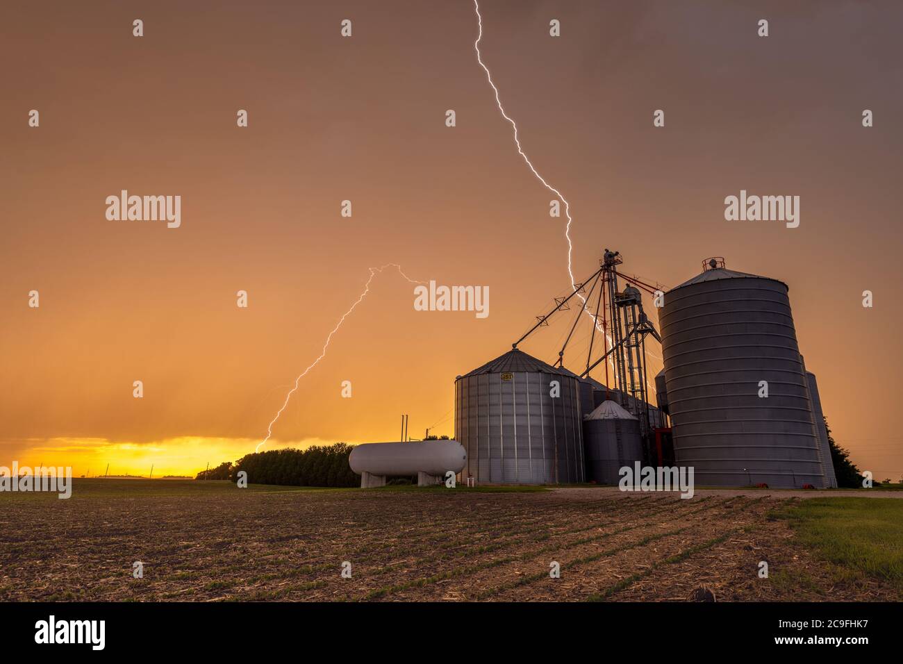 Fulmini che sparano da un cielo arancione al tramonto con un impianto di lavorazione del grano in primo piano. Foto Stock