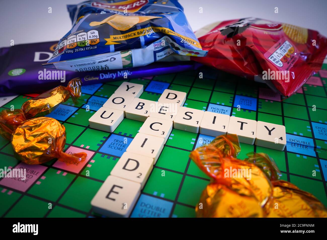 Primo piano di Scrabble lettere di gioco parole di ortografia: Obesità si decide, a bordo con spuntini cibo spazzatura. Gioco della vita. Foto Stock