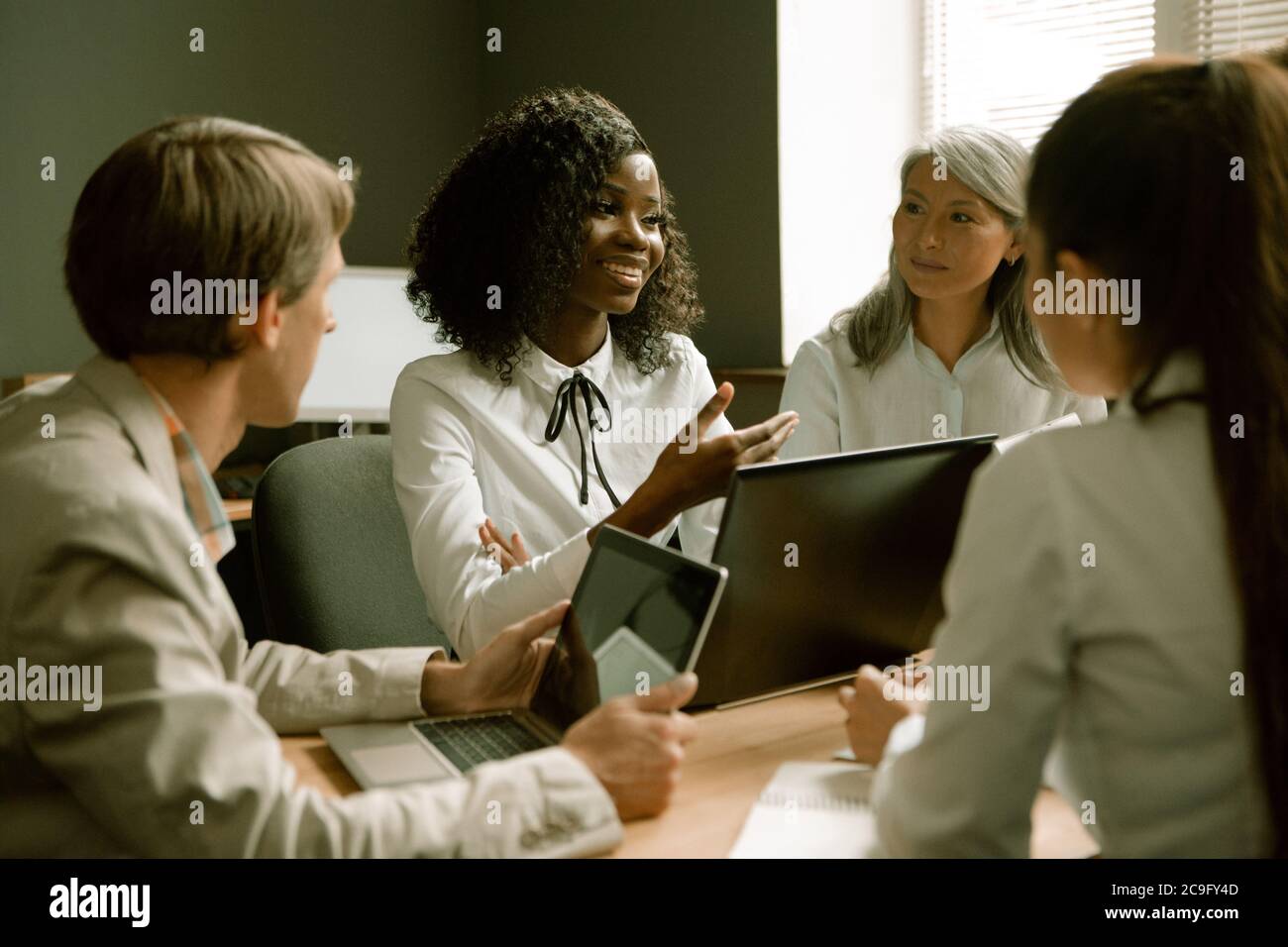 La gente felice di affari chiacchiera durante la riunione di affari in ufficio. Focalizzazione selettiva sulle donne africane che condividono idee con i colleghi. Immagine colorata Foto Stock