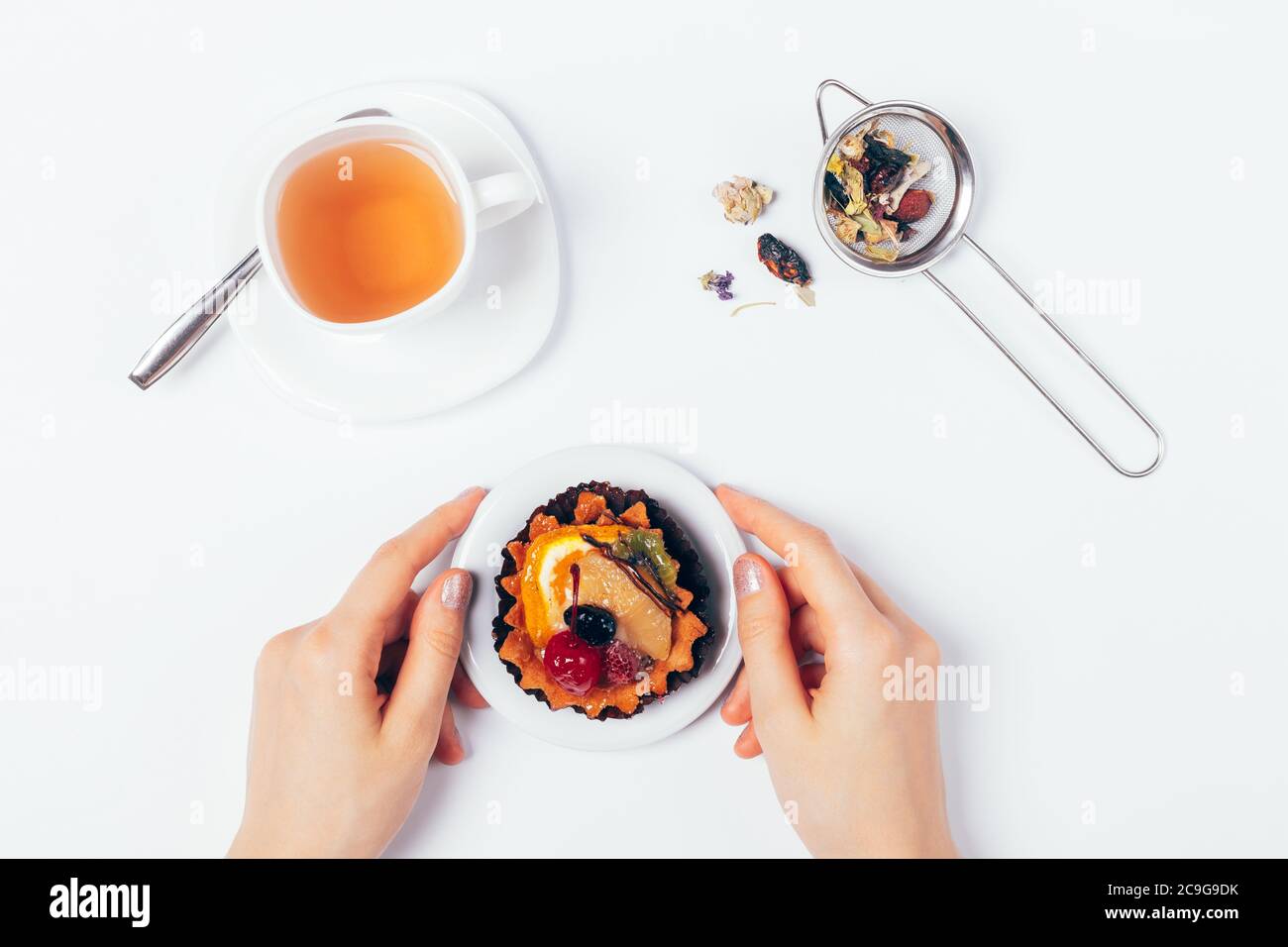 Le mani femminili tengono la torta del dessert vicino alla tazza del tè fresco e del filtro con le erbe secche e i fiori sul tavolo bianco, vista dall'alto. Foto Stock