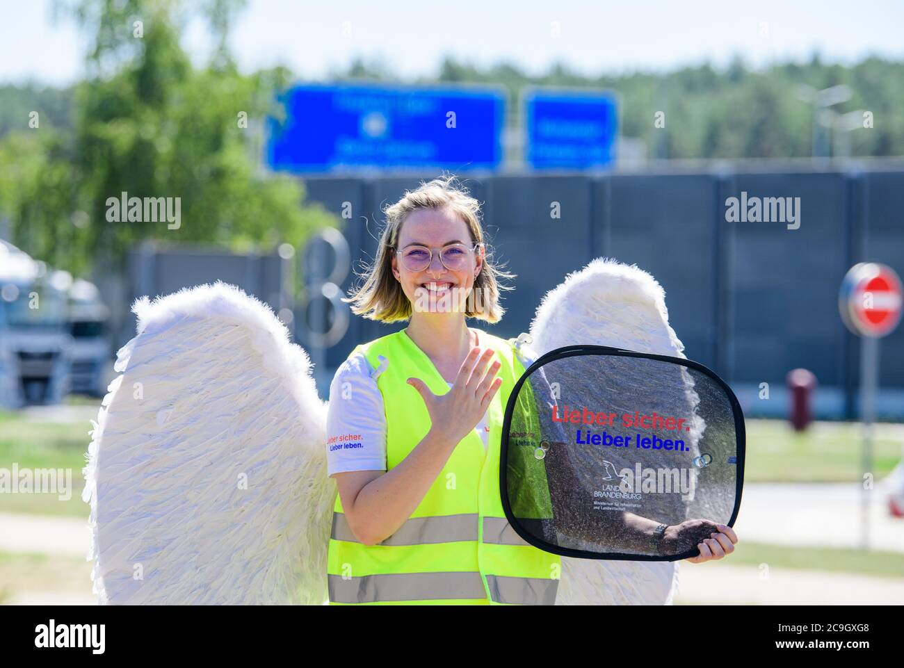 31 luglio 2020, Brandeburgo, Michendorf: Emelie, vestito da angelo custode Jo, si erge sui terreni della fermata di sosta Michendorf-Nord e tiene un distributore di ombrellone con l'iscrizione 'Lieber sicher. Meglio vivere.' nelle sue mani. Durante la campagna di sicurezza stradale con lo stesso nome, un tour di tutte le fermate di sosta del Brandeburgo è quello di attirare l'attenzione sui pericoli del traffico stradale, soprattutto durante i viaggi di vacanza. A tal fine, verrà allestito uno stand informativo della polizia, un modello di corsia di emergenza, un'auto di bobby e una pista sportiva, uno stand dell'ADAC per i test di reazione e una ruota di fortuna. Th Foto Stock