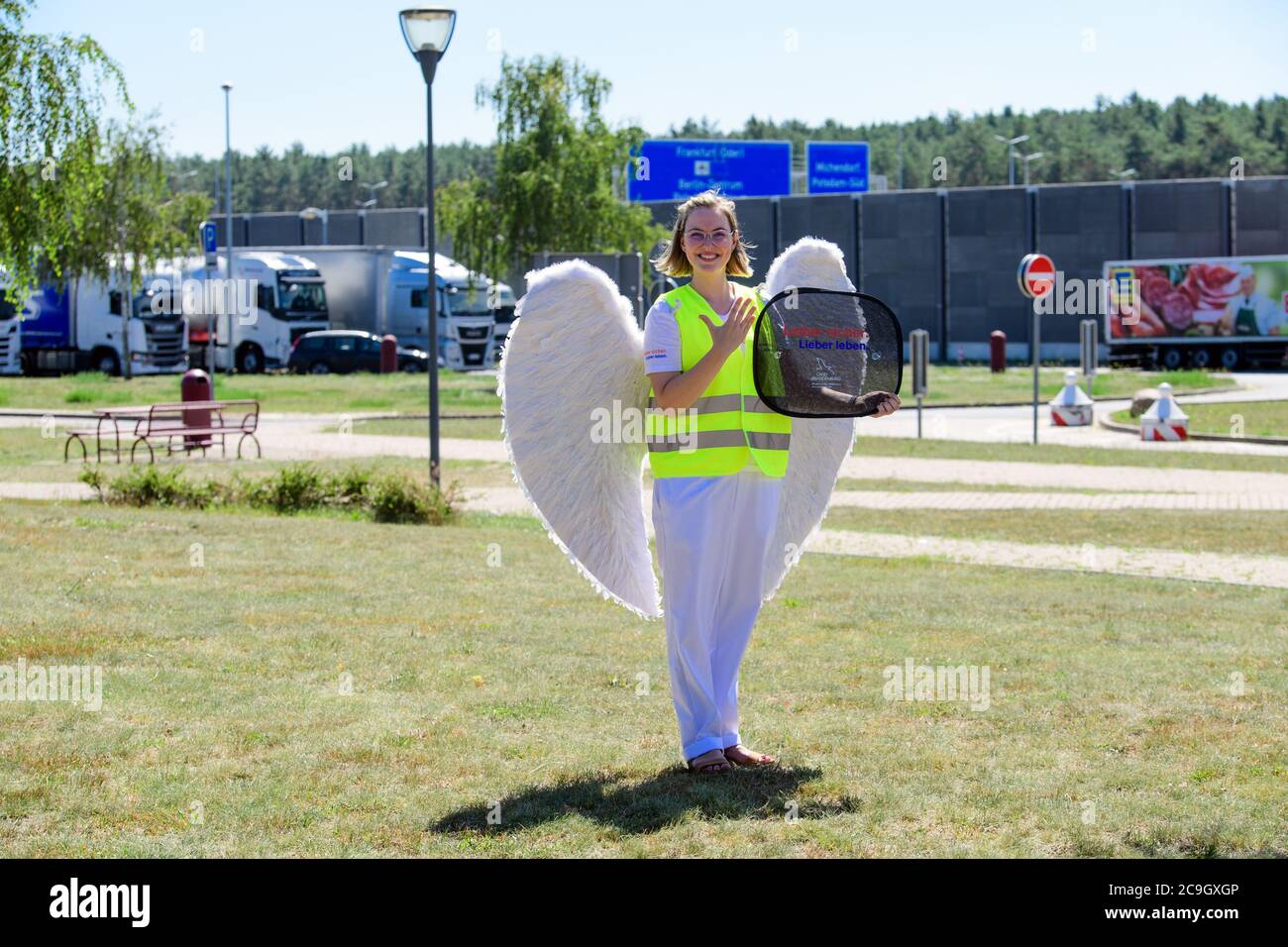 31 luglio 2020, Brandeburgo, Michendorf: Emelie, vestito da angelo custode Jo, si erge sui terreni della fermata di sosta Michendorf-Nord e tiene un distributore di ombrellone con l'iscrizione 'Lieber sicher. Meglio vivere.' nelle sue mani. Durante la campagna di sicurezza stradale con lo stesso nome, un tour di tutte le fermate di sosta del Brandeburgo è quello di attirare l'attenzione sui pericoli del traffico stradale, soprattutto durante i viaggi di vacanza. A tal fine, verrà allestito uno stand informativo della polizia, un modello di corsia di emergenza, un'auto di bobby e una pista sportiva, uno stand dell'ADAC per i test di reazione e una ruota di fortuna. Th Foto Stock