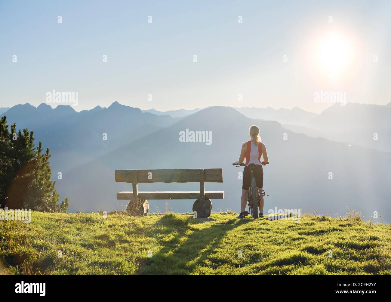 Il biker di montagna si trova accanto a una panchina di legno e si affaccia sul vasto paesaggio di montagna inondato di luce, Patscherkofel, Patsch, Tirolo, Austria Foto Stock