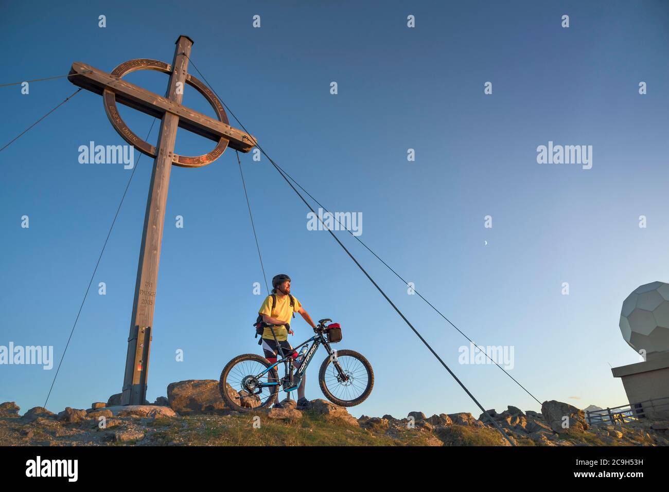 Il biker di montagna si erge con e-MTB accanto alla croce di cima del Patscherkofel nella calda luce della sera, Patscherkofel, Patsch, Tirolo, Austria Foto Stock