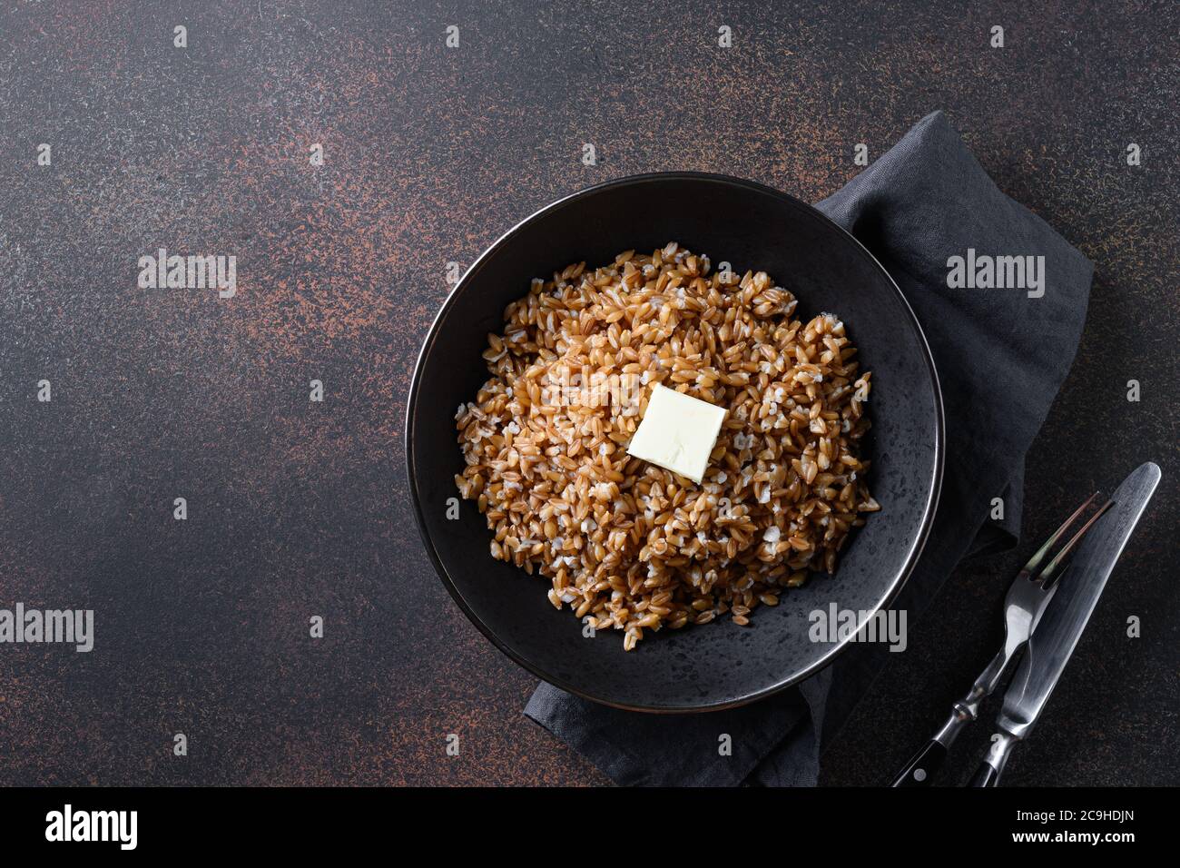 Farro di cereali interi cotti con burro in ciotola nera su fondo marrone scuro. Vista dall'alto. Foto Stock