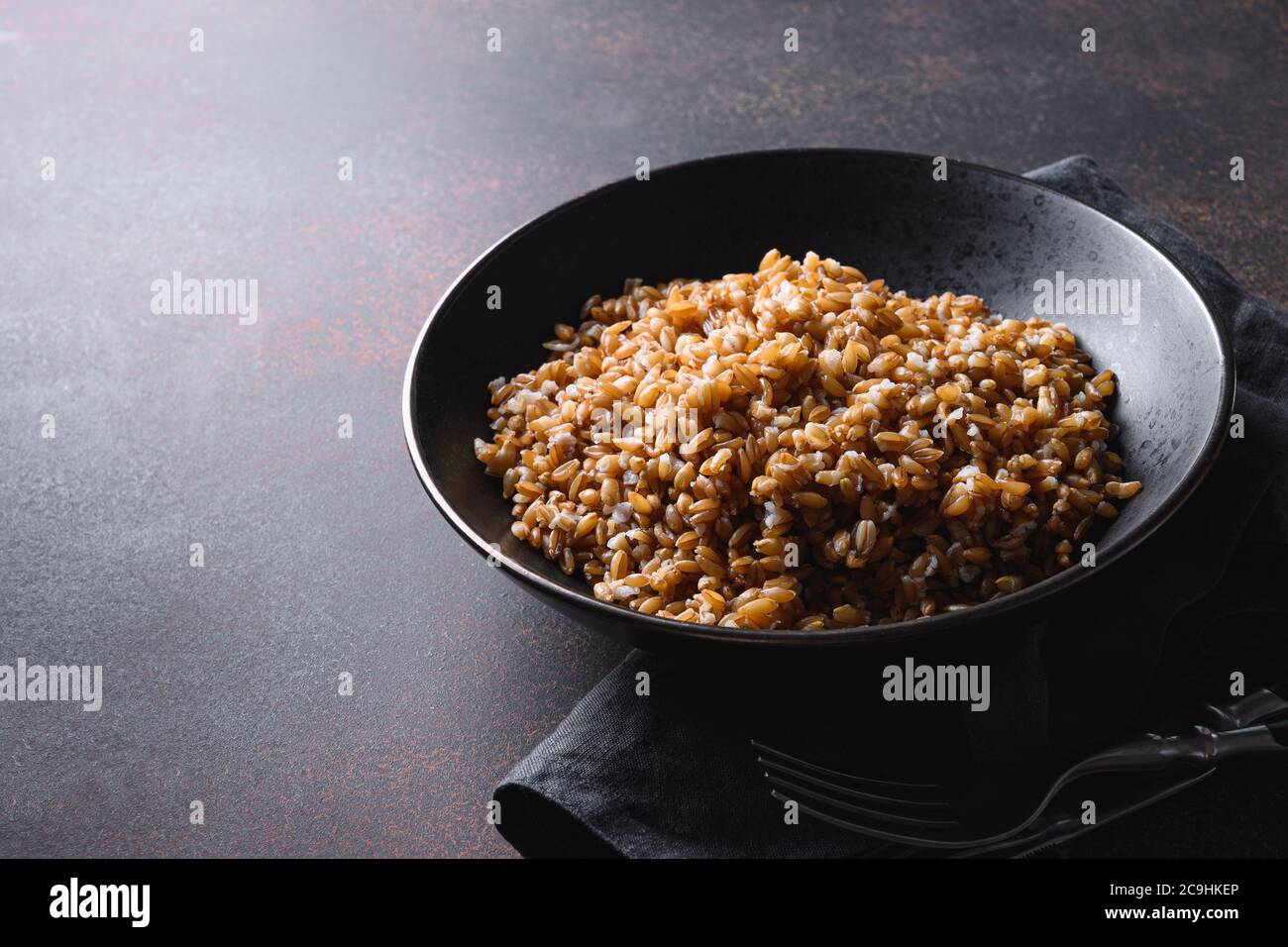 Farro di cereali interi cotti in ciotola nera su fondo marrone scuro. Primo piano. Foto Stock