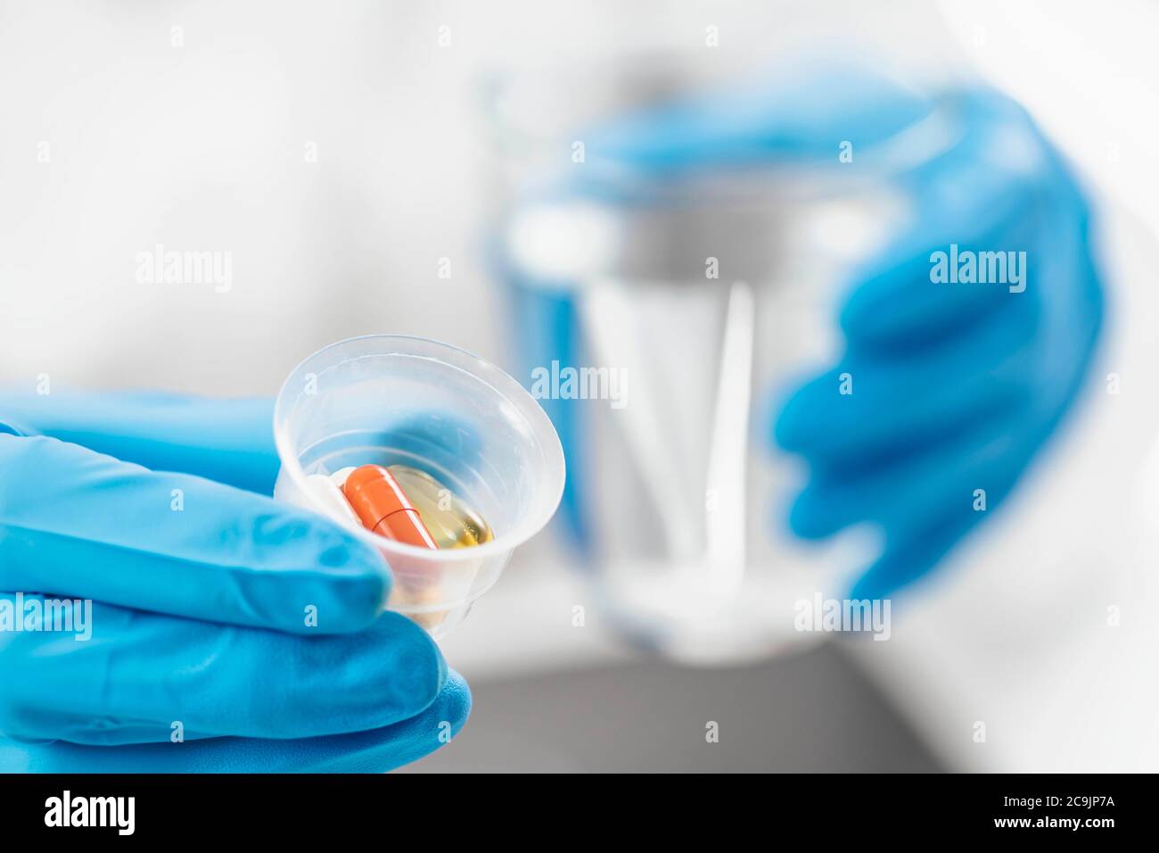 La mano del medico femminile in guanto blu che tiene la tazza graduata della medicina piena di pillole e del bicchiere d'acqua. Foto Stock