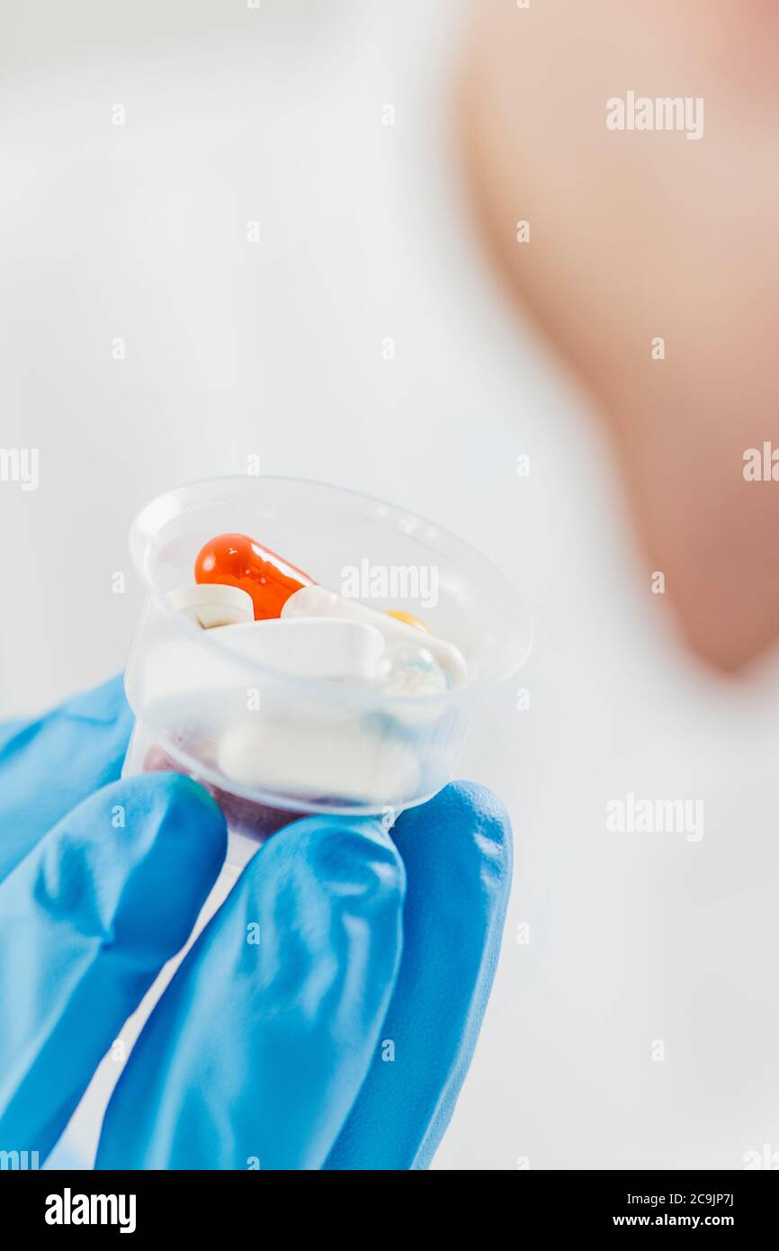 La mano del medico femminile in guanto blu che tiene i bicchieri graduati della medicina pieni di pillole. Copia spazio e sfondo bianco. Foto Stock