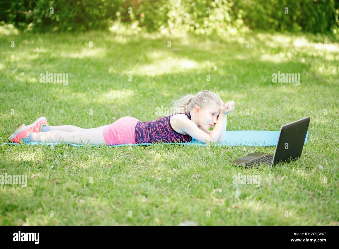 Bambino stanco annoiato che fa l'allenamento di sport all'aperto in linea. Corso di video yoga su Internet. Divertente allenamento sonnolento per bambini nel cortile di casa con il computer portatile. Foto Stock