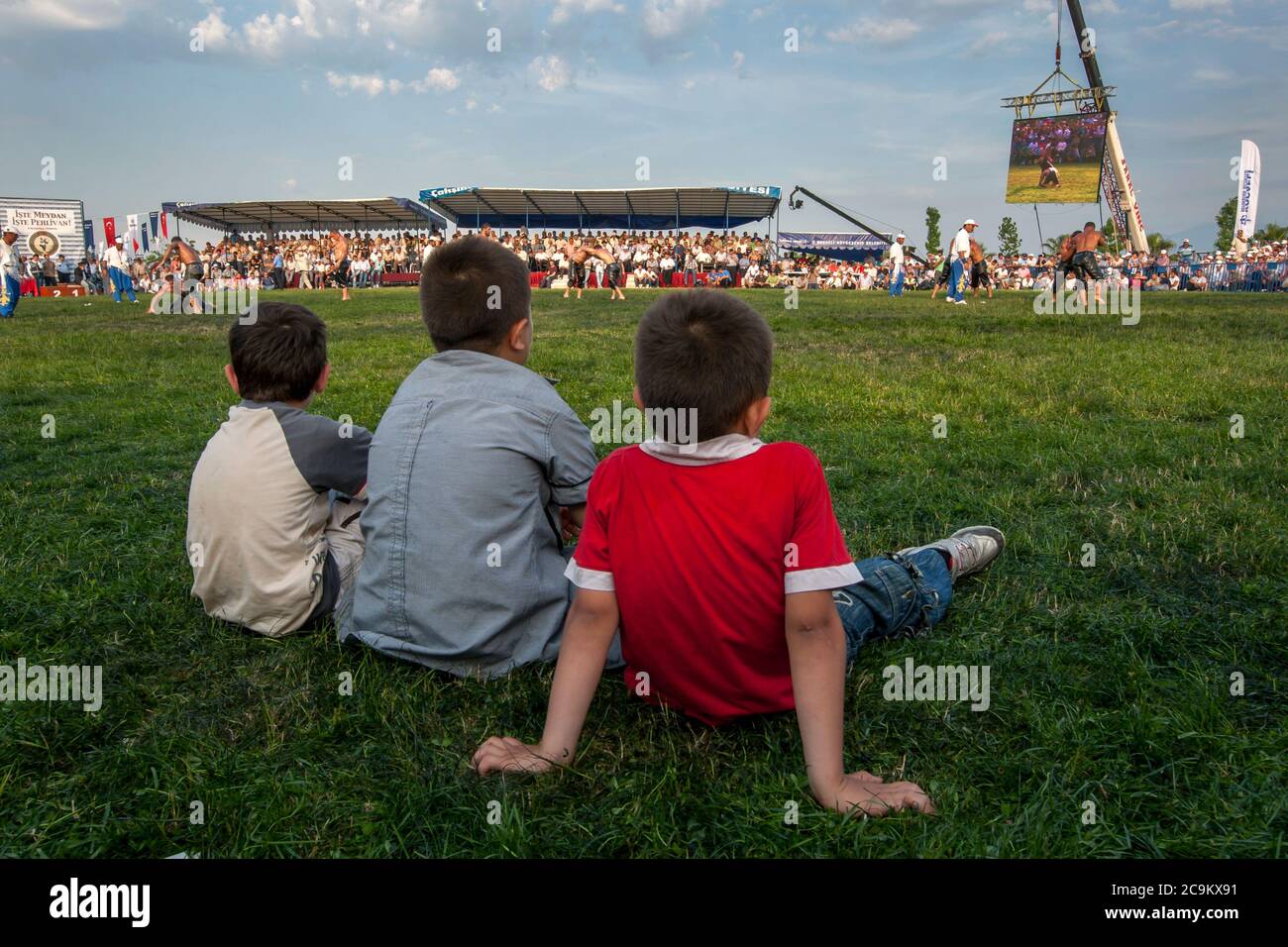 Tre ragazzi si siedono nell'arena della competizione per assistere alla battaglia dei lottatori di pesi pesanti al festival Izmit Turkish Oil Wrestling in Turchia. Foto Stock