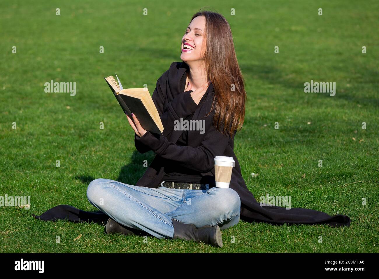 bruna ragazza si siede a croce su un prato verde e ride gioiosamente mentre legge un libro nero hardback con un bicchiere di carta da caffè eco. Foto Stock