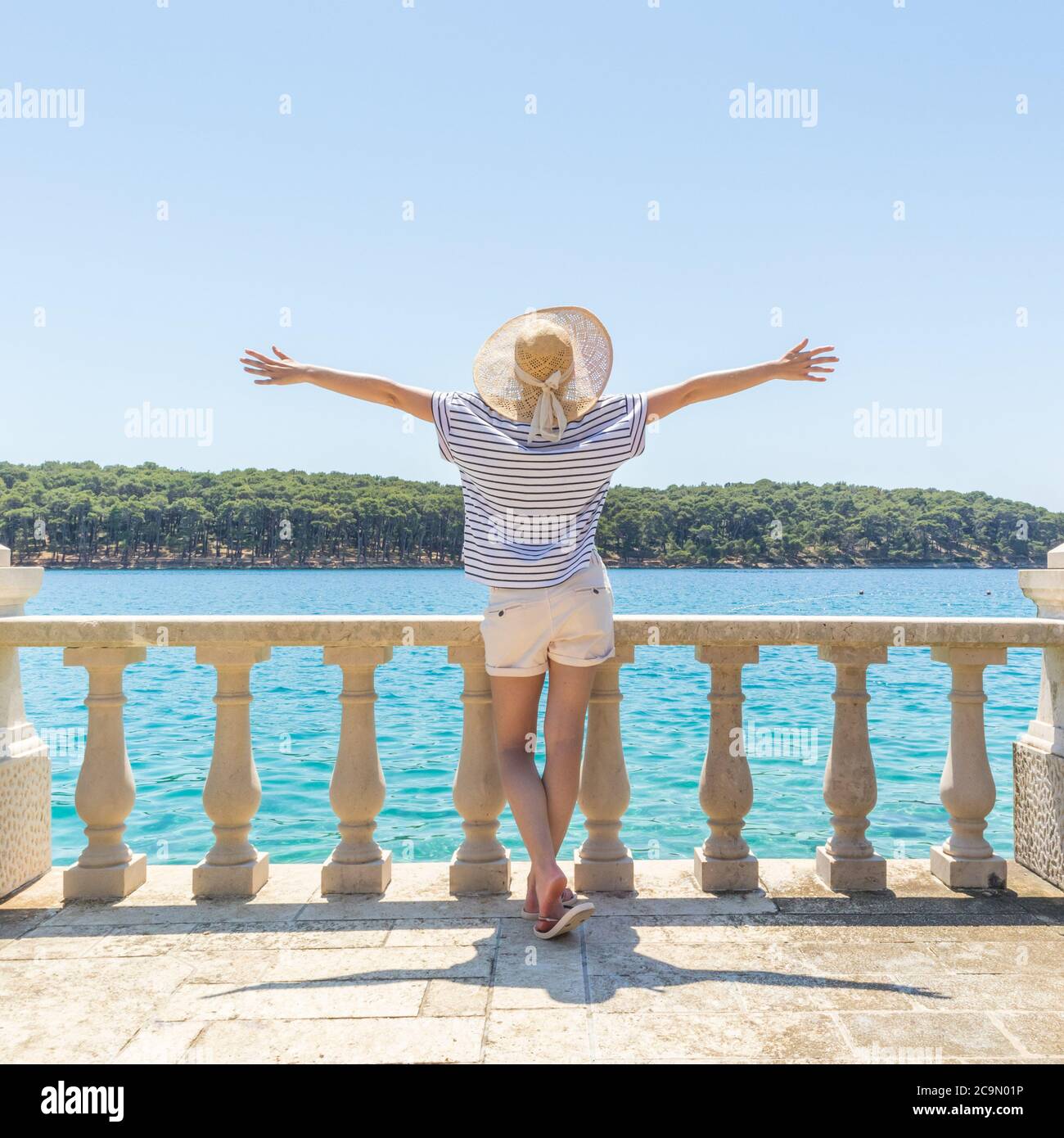 Vista posteriore di donna felice in vacanza, indossando cappello d'estate paglia, in piedi su elegante balcone di pietra di lusso della villa costiera, rilassante, braccia rischiate Foto Stock