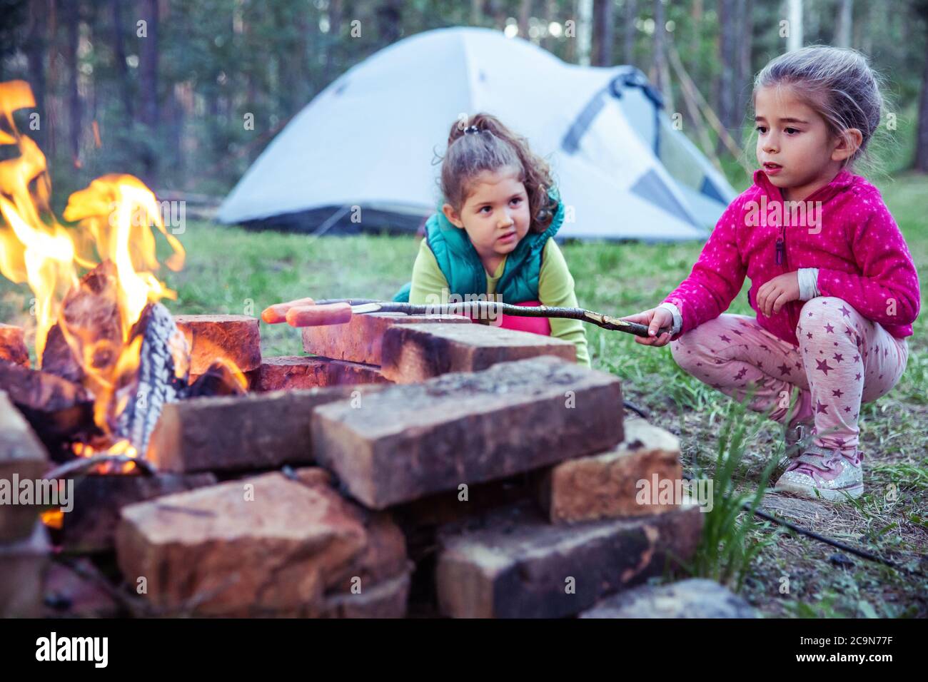 Due bambini piccoli preparano il cibo al falò, con tenda da campeggio sullo sfondo. Foto Stock
