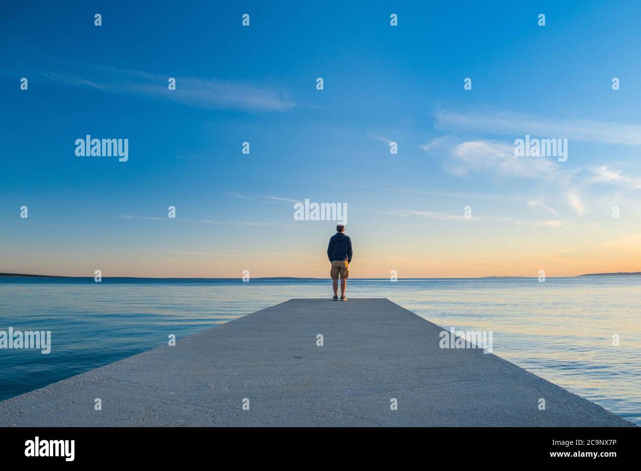 Uomo in felpa con cappuccio e pantaloni da carico corti in piedi sul molo e guardando all'orizzonte del mare, isola d Pag, mare Adriatico, Croazia Foto Stock