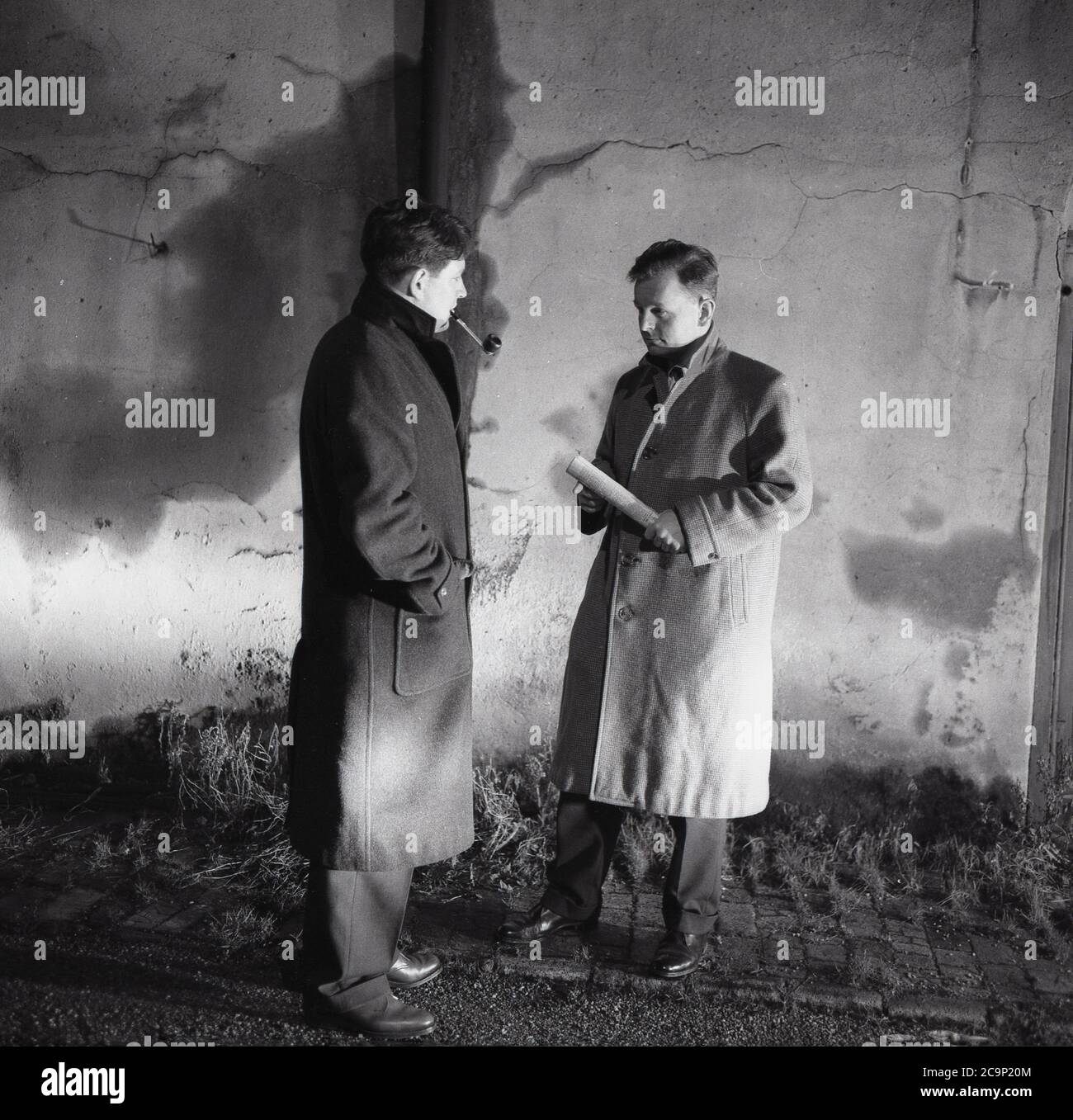 Anni Sessanta, storico, sera e accanto ad un alto muro, due uomini che indossano soprabiti che hanno un incontro segreto o clandestino, con uno degli uomini che fuma un tubo e l'altro che tiene un rotolo di documenti, Londra, Inghilterra, Regno Unito. Foto Stock