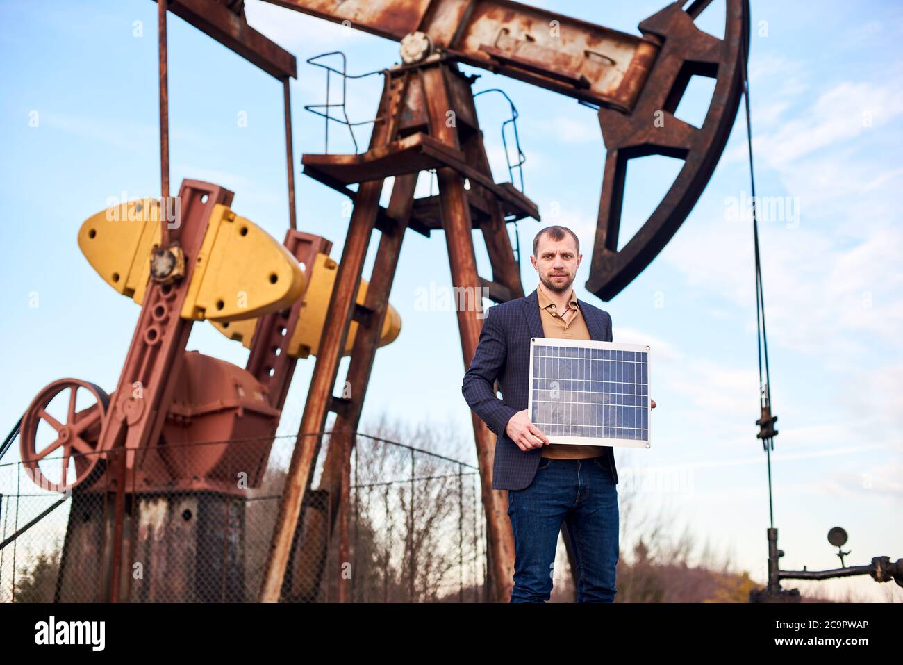 Ritratto orizzontale di un uomo d'affari che indossa un cappotto sportivo, jeans che tengono mini batteria solare, olio pompa jack dietro di lui, concetto di fonte alternativa di energia Foto Stock