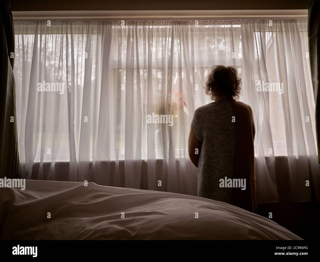 Donna anziana vulnerabile che si autoisola e scherma da Coronavirus / Covid-19 pandemia guardando fuori la sua finestra di isolamento COVID blocco camera da letto. REGNO UNITO Foto Stock