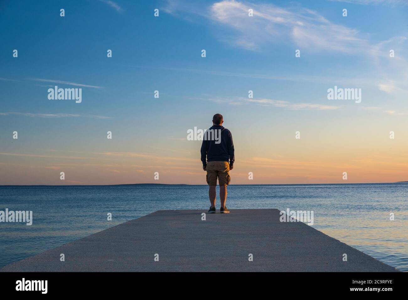 Uomo in felpa con cappuccio e pantaloni da carico corti in piedi sul molo e guardando all'orizzonte del mare, isola d Pag, mare Adriatico, Croazia Foto Stock