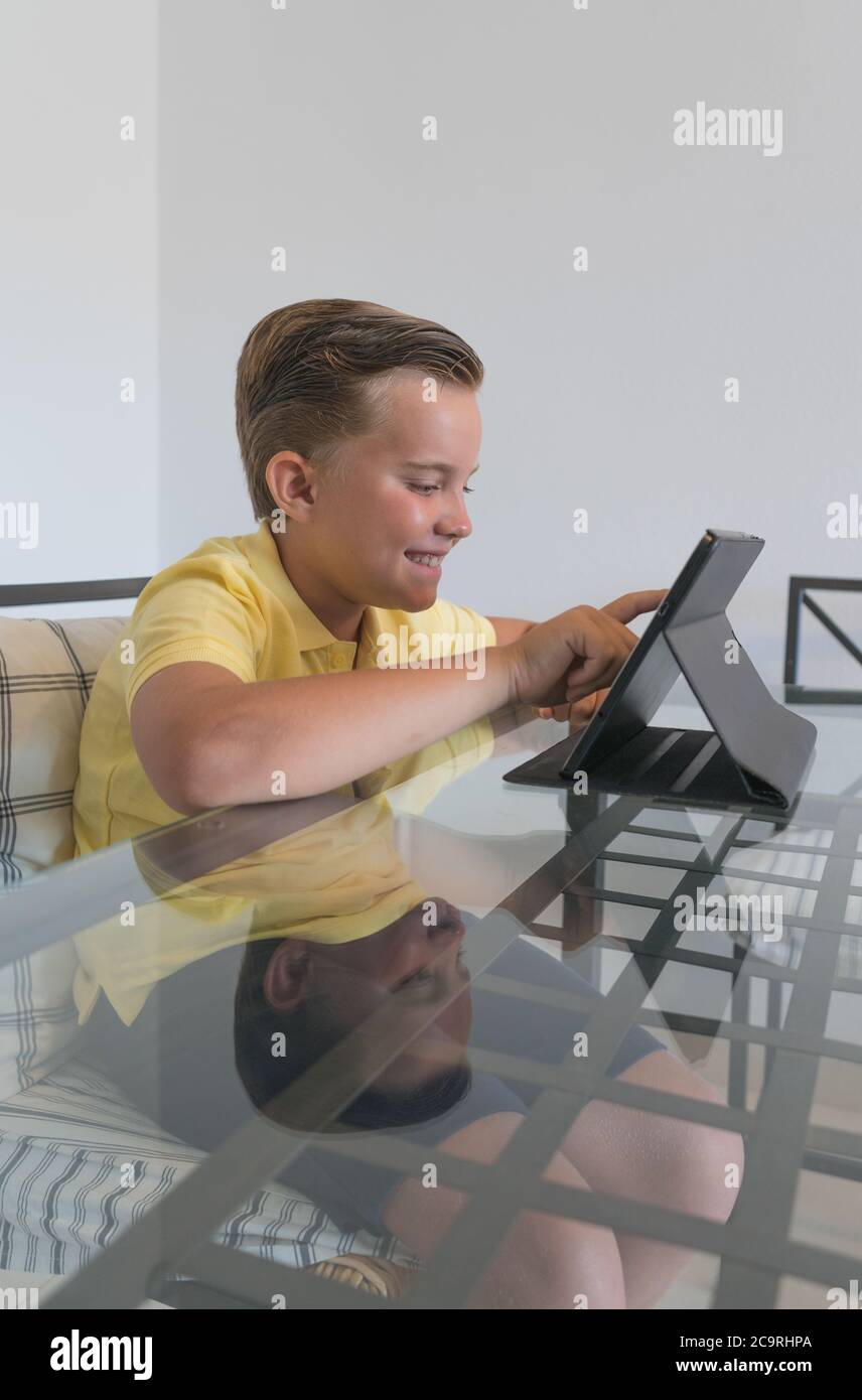 Ragazzo che parla con il compagno di classe durante la videochiamata sul tablet e discute di lavoro mentre si siede al tavolo in una stanza moderna e luminosa. Foto Stock