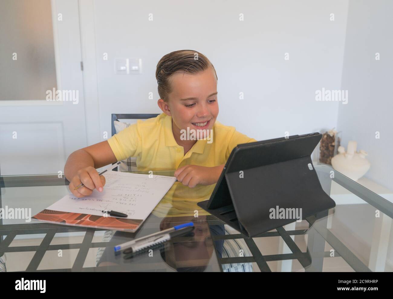 Ragazzo che parla con il compagno di classe durante la videochiamata sul tablet e discute di lavoro mentre si siede al tavolo in una stanza moderna e luminosa. Foto Stock