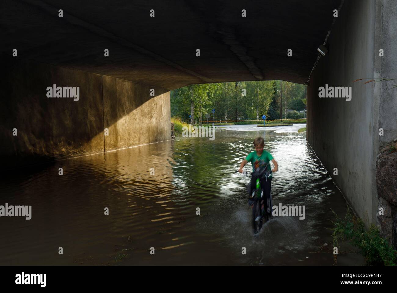 Giovane ragazzo caucasico pedalando attraverso il sottopassaggio stradale allagato dopo il deflusso a Estate, Finlandia Foto Stock