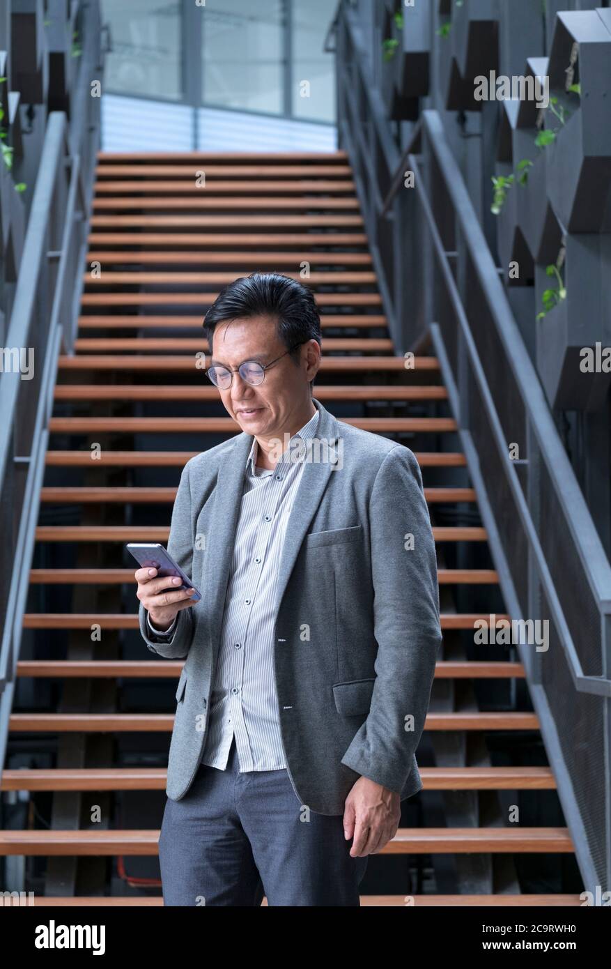 Uomo d'affari cinese in piedi sotto la scala e guardando il cellulare in mano. Foto Stock