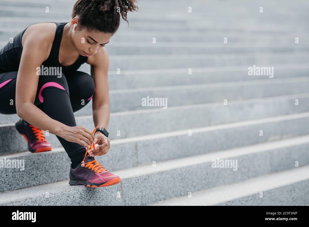 Runner si prepara per la formazione. Ragazza afroamericana con cuffie wireless e lacci per il fitness tracker sulle sneakers Foto Stock