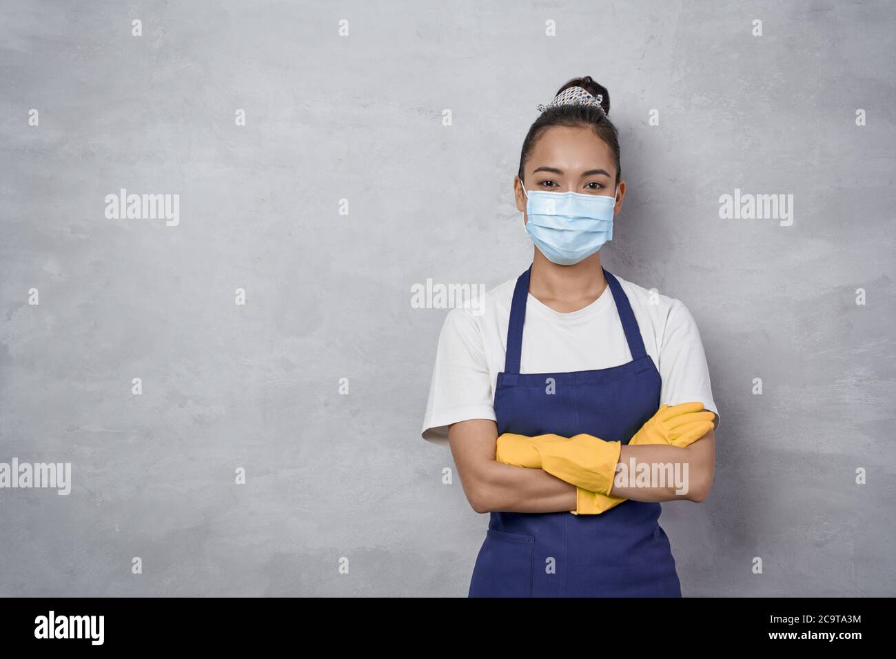 Donna di pulizia sicura che indossa guanti in gomma e maschera medica protettiva per il viso tenendo le braccia incrociate, guardando la fotocamera mentre si trova contro la parete grigia. Servizi di pulizia durante la covid 19 pandemia Foto Stock