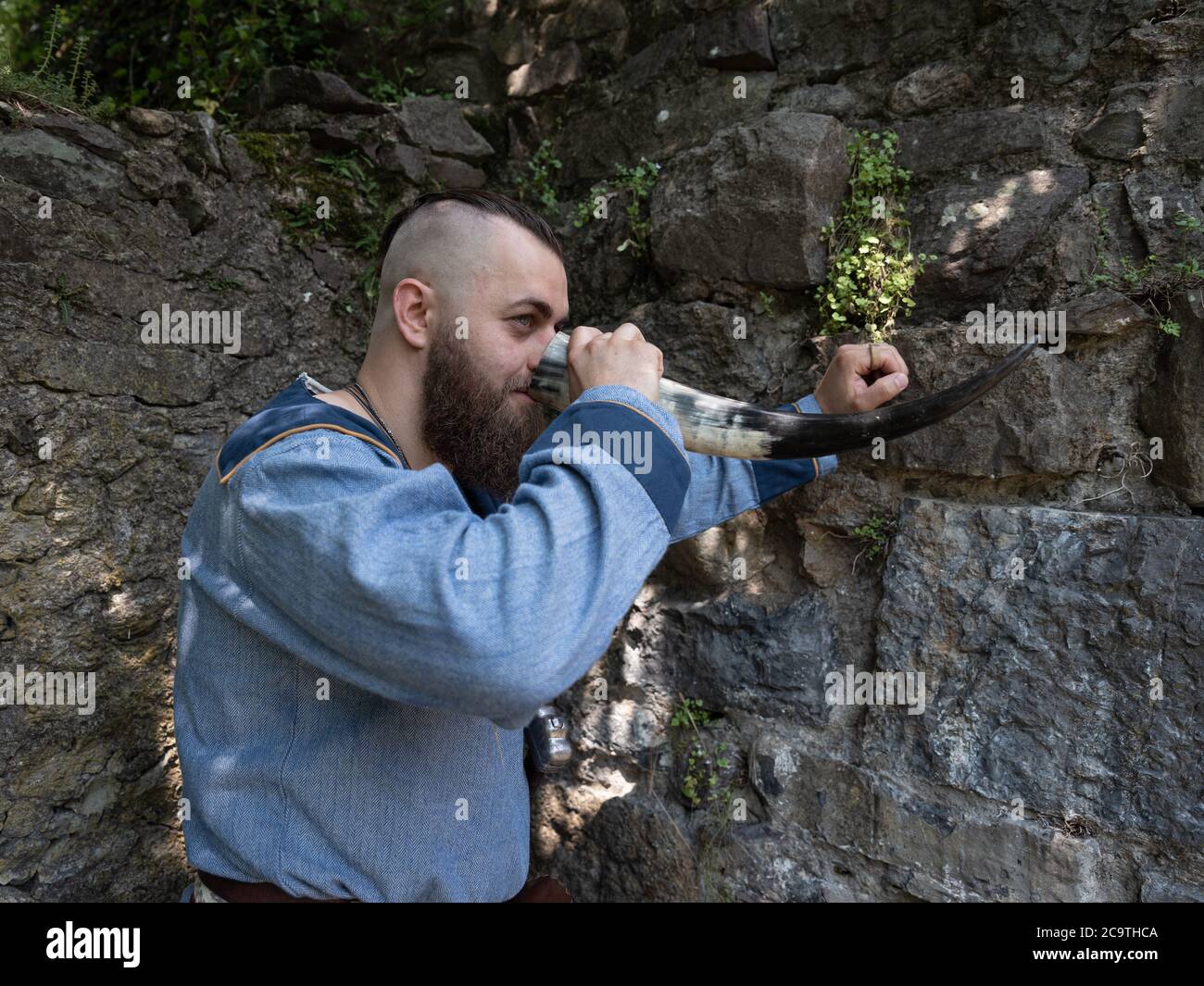 Un guerriero vichingo beve sidro da un corno appoggiato su un muro di pietra, immagine dell'uomo in abiti medievali Foto Stock