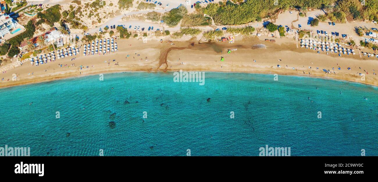 Vista aerea dall'alto della bellissima spiaggia di Coral a Paphos con acque marine azzurre, Cipro. Costa di sabbia con ombrelloni, lettini, persone e acqua di mare limpida. Foto Stock