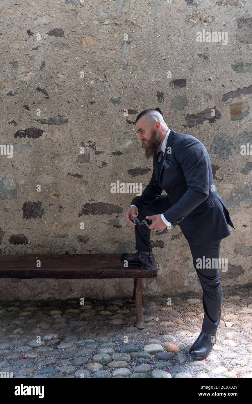 L'uomo in abiti eleganti poggia un piede su una panchina di legno in un piccolo villaggio Foto Stock