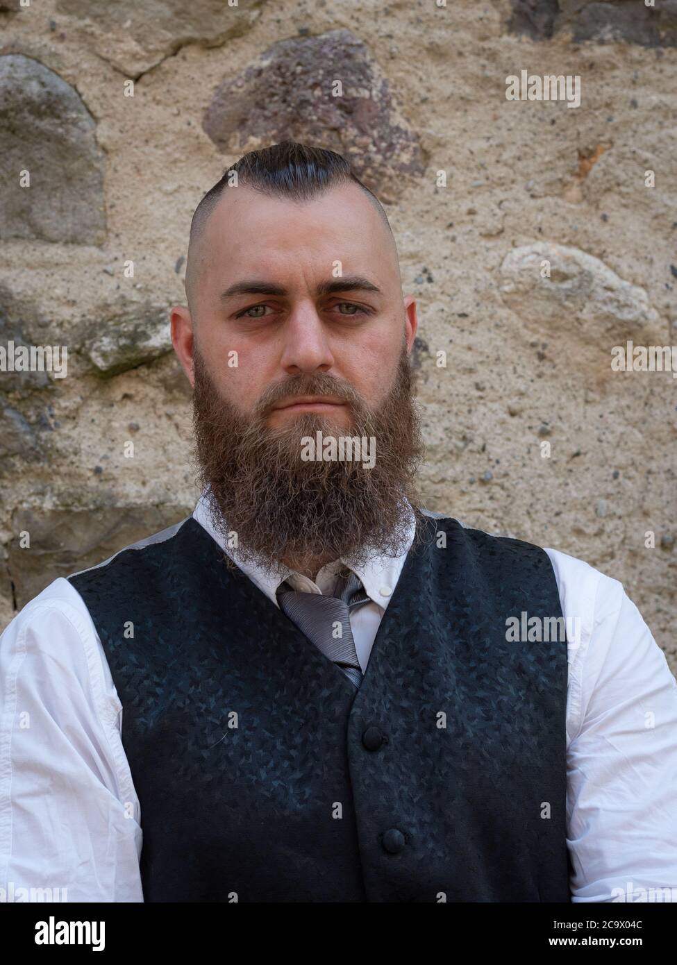 L'uomo con barba lunga indossa un abito elegante e scuro che si posa davanti a un muro di pietra Foto Stock
